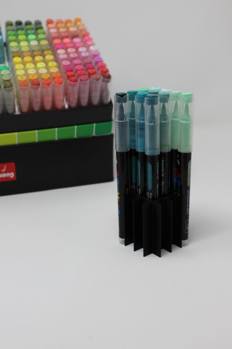 A collection of colorful art markers with clear caps, arranged in a black organizer. The markers are in various shades, including blue, green, pink, and yellow. A second set of markers is visible in the background.