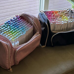 May include: Two open art supply bags filled with colorful markers. One bag is pink with a clear top, and the other is black. The markers are arranged in a rainbow of colors, perfect for artists and crafters.