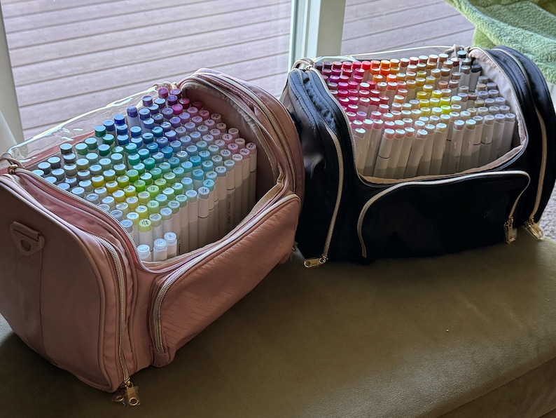May include: Two open art supply bags filled with colorful markers. One bag is pink with a clear top, and the other is black. The markers are arranged in a rainbow of colors, perfect for artists and crafters.
