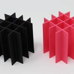 Two geometric cube-shaped organizers, one black and one bright pink, stand side-by-side. Each is constructed from intersecting vertical and horizontal panels, creating a grid-like structure. The design is modern and minimalist.