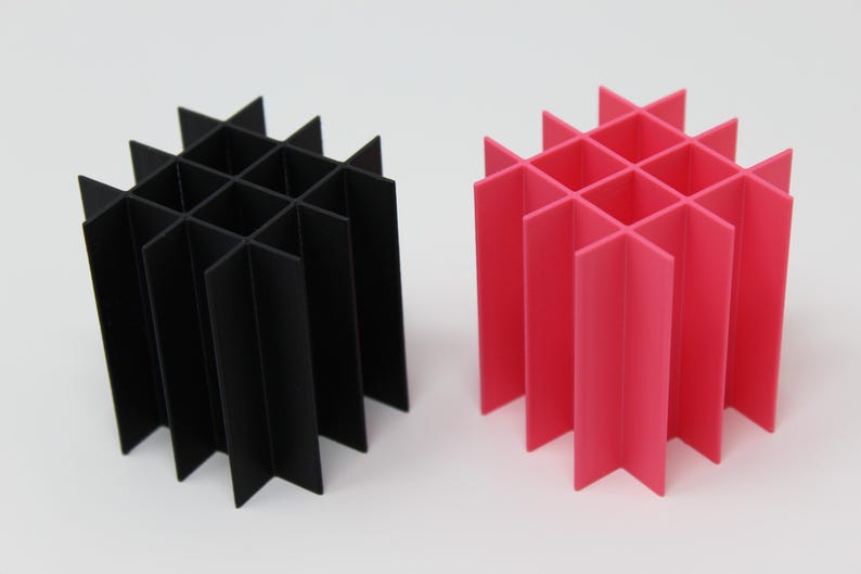 Two geometric cube-shaped organizers, one black and one bright pink, stand side-by-side. Each is constructed from intersecting vertical and horizontal panels, creating a grid-like structure. The design is modern and minimalist.