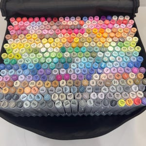 May include: A large collection of colorful art markers arranged in a black carrying case. The markers are in a wide array of colors, including pinks, blues, greens, yellows, and grays. The caps of the markers are color-coded.