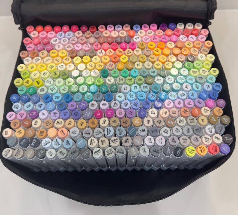 May include: A large collection of colorful art markers arranged in a black carrying case. The markers are in a wide array of colors, including pinks, blues, greens, yellows, and grays. The caps of the markers are color-coded.