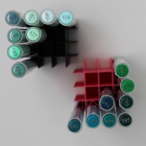 A collection of colorful art markers in shades of green and blue, organized in black and pink plastic holders. The markers are arranged in rows, with the numbers visible on the caps. The background is plain white.