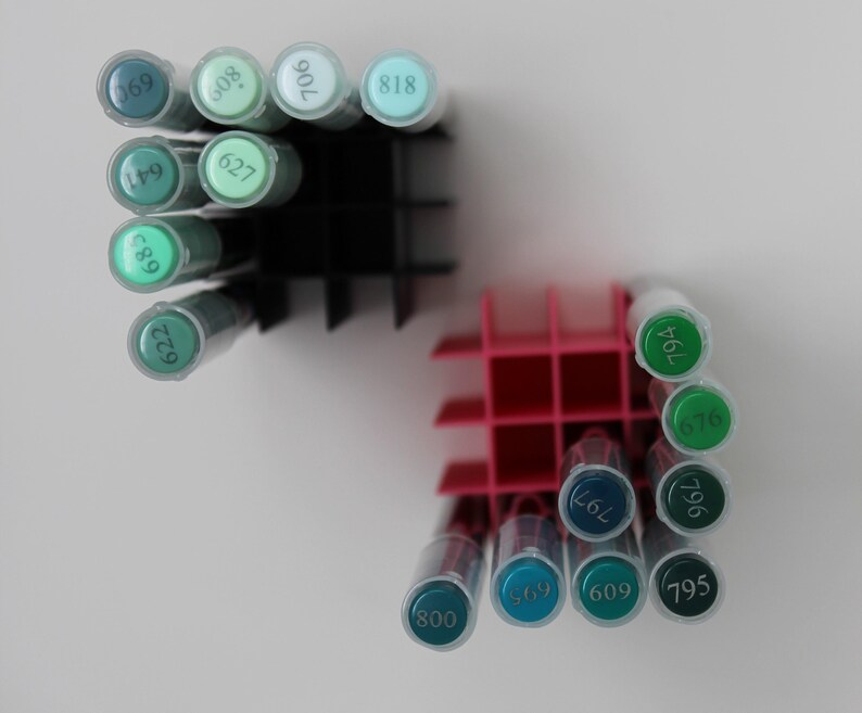 A collection of colorful art markers in shades of green and blue, organized in black and pink plastic holders. The markers are arranged in rows, with the numbers visible on the caps. The background is plain white.