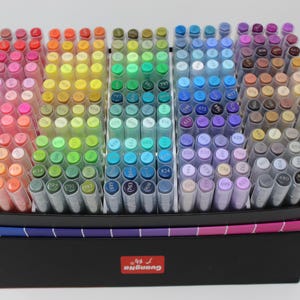 A collection of colorful art markers arranged in a black box. The markers are organized by color, ranging from reds and oranges to blues, greens, and purples. The box has a rainbow-colored trim and a red logo.