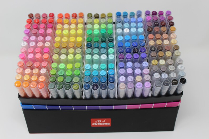 A collection of colorful art markers arranged in a black box. The markers are organized by color, ranging from reds and oranges to blues, greens, and purples. The box has a rainbow-colored trim and a red logo.