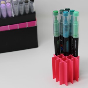 A collection of art markers with clear caps and black barrels, some with green and blue accents, are displayed in a bright pink organizer. A black box with a color chart and the brand name "Guangna" is in the background.