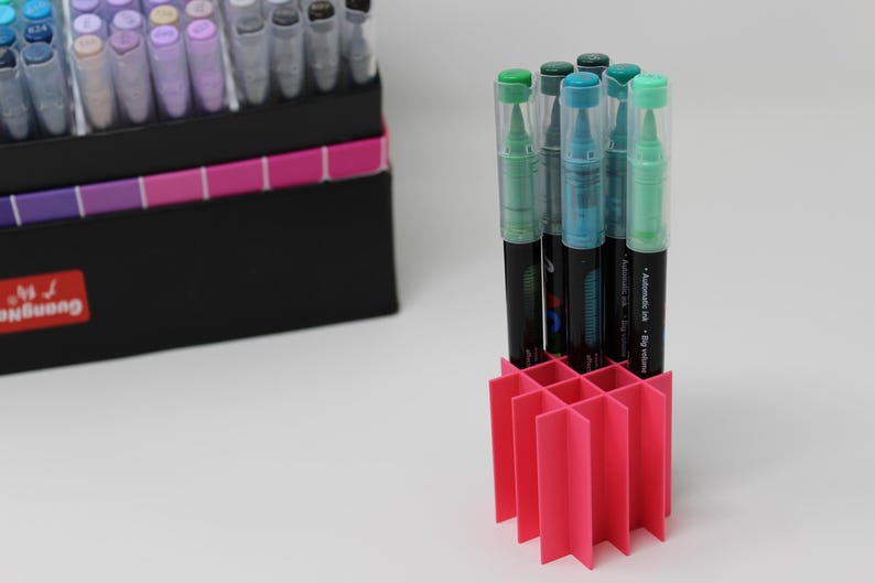 A collection of art markers with clear caps and black barrels, some with green and blue accents, are displayed in a bright pink organizer. A black box with a color chart and the brand name "Guangna" is in the background.