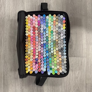 May include: A black carrying case filled with a large collection of colorful art markers. The markers are arranged in rows, showcasing a wide spectrum of colors, including shades of red, blue, green, yellow, and purple. The case has a handle for easy transport.