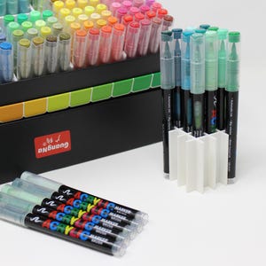 A collection of colorful acrylic markers in a black box with a rainbow-colored divider. Several markers are in a white holder, and a few are laid out. The markers have clear caps and black barrels with colorful text.