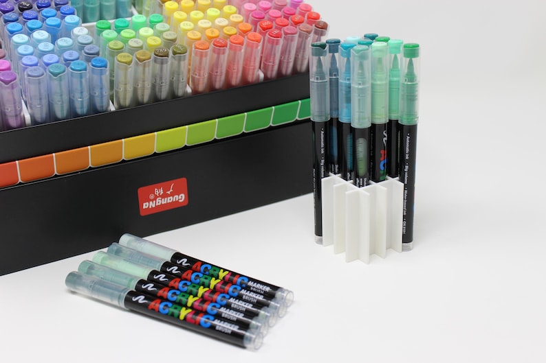 A collection of colorful acrylic markers in a black box with a rainbow-colored divider. Several markers are in a white holder, and a few are laid out. The markers have clear caps and black barrels with colorful text.
