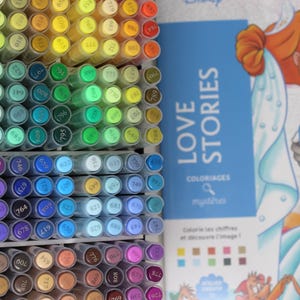 A collection of colorful art markers arranged in a grid, with a book titled "LOVE STORIES" featuring a Disney illustration. The markers are in various shades, including green, blue, purple, and yellow. The book cover shows a couple kissing.