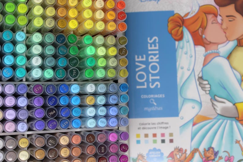 A collection of colorful art markers arranged in a grid, with a book titled "LOVE STORIES" featuring a Disney illustration. The markers are in various shades, including green, blue, purple, and yellow. The book cover shows a couple kissing.