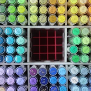 A close-up view of a collection of colorful art markers, neatly organized in a white storage unit. The markers are arranged by color, with shades of pink, orange, yellow, green, blue, and purple. Each marker has a number on its cap.