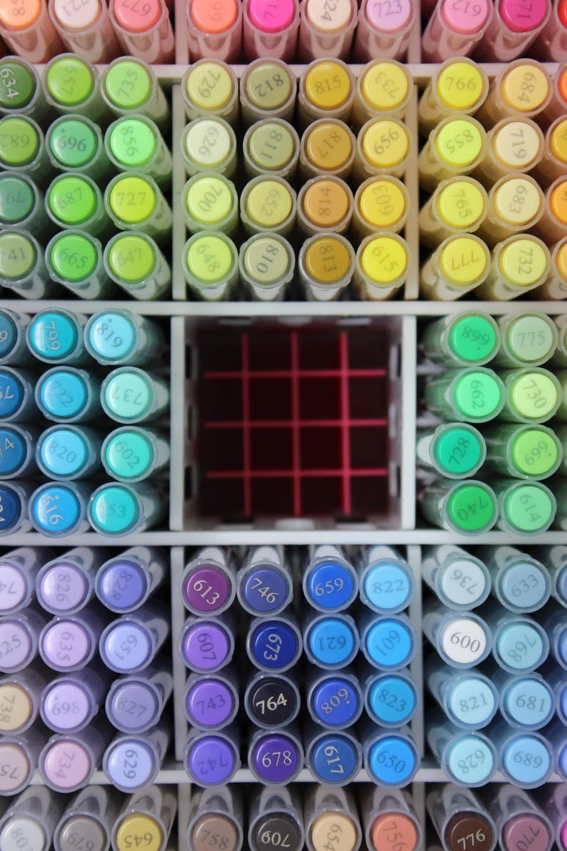 A close-up view of a collection of colorful art markers, neatly organized in a white storage unit. The markers are arranged by color, with shades of pink, orange, yellow, green, blue, and purple. Each marker has a number on its cap.