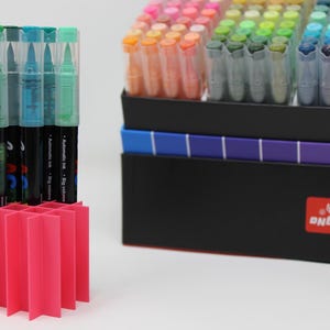 A collection of colorful art markers with clear caps, displayed in a bright pink organizer. The markers are in a black box with a color gradient and a red GuangNa label.