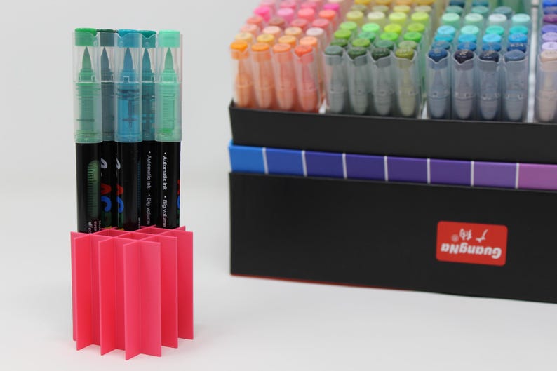 A collection of colorful art markers with clear caps, displayed in a bright pink organizer. The markers are in a black box with a color gradient and a red GuangNa label.