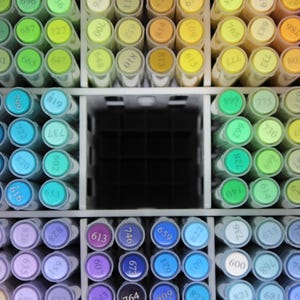 A collection of colorful art markers arranged in a white organizer. The markers are in various shades of green, blue, purple, yellow, and orange. Each marker has a number on its cap, indicating the color.