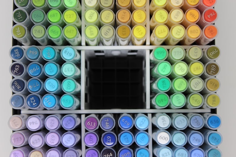 A collection of colorful art markers arranged in a white organizer. The markers are in various shades of green, blue, purple, yellow, and orange. Each marker has a number on its cap, indicating the color.
