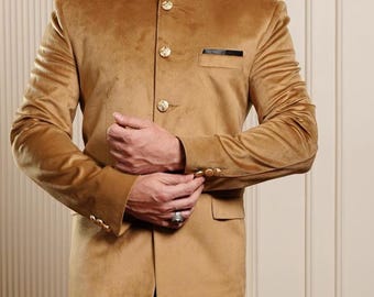 Puede incluir: Un blazer de terciopelo color canela con botones dorados y cuello mandarín se combina con pantalones de vestir negros. La chaqueta tiene un pequeño bolsillo y mangas con puños. El conjunto se completa con mocasines de charol negros.