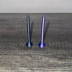 May include: Two metal golf tees standing upright. The left tee has a dark, iridescent blue-grey finish, while the right tee features a vibrant, polished purple metallic finish. Both items have a pointed tip and a flat, circular base for stability.