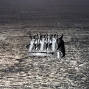 Op de afbeelding: Een zilverkleurige, 3D-geprinte plastic clip met een rij van vier halfronde, aan de bovenkant open cilindrische houders die naast elkaar zijn geplaatst. Het object heeft een getextureerde, matte afwerking en een gebogen, veerachtige basis.