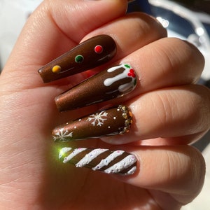 May include: Coffin-shaped, brown nail art with festive gingerbread designs. Some nails feature white icing, colorful candies, and snowflake accents. Others have a stripe design with glitter.