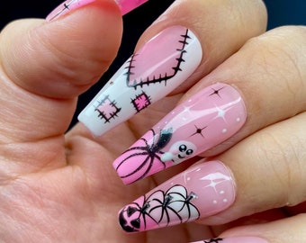 May include: Pink and white long coffin-shaped fake nails with Halloween-themed designs. The designs include a ghost, pumpkins, and spiderwebs.