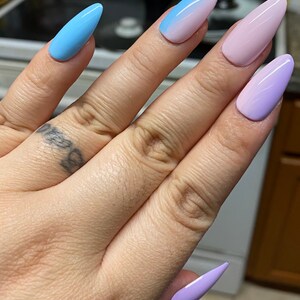 May include: A hand model wearing five long, stiletto-shaped false nails with ombre nail polish. The nails are painted in shades of blue, pink, and purple.