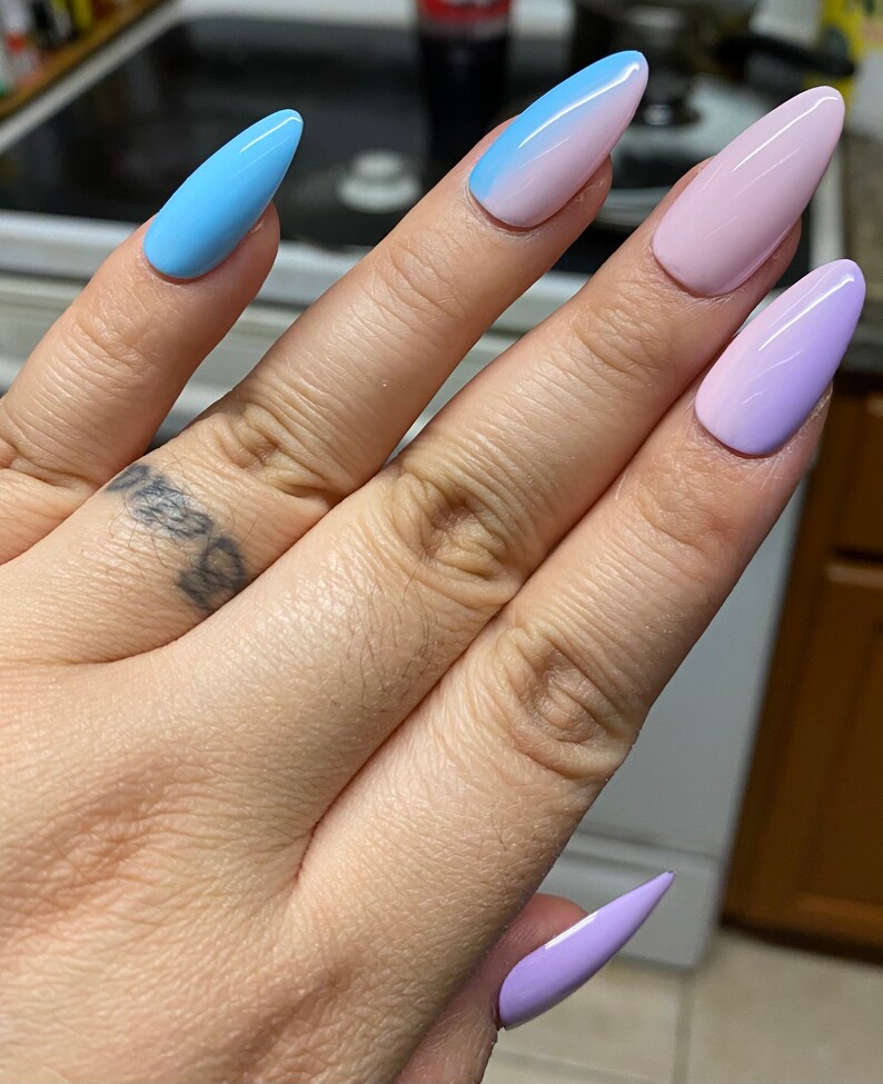 May include: A hand model wearing five long, stiletto-shaped false nails with ombre nail polish. The nails are painted in shades of blue, pink, and purple.