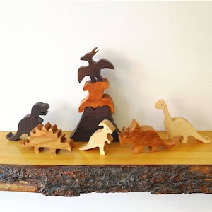 Peut inclure: Un ensemble de jouets en bois de dinosaures, comprenant un Tyrannosaurus Rex, un Stégosaure, un Tricératops, un Ptérodactyle et un volcan. Les jouets sont peints dans des tons de brun, orange et noir.