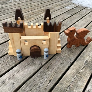 May include: A wooden castle toy set featuring two knights and a dragon. The castle is crafted from natural wood with brown accents. The knights are painted grey with blue shields. The dragon is painted red.