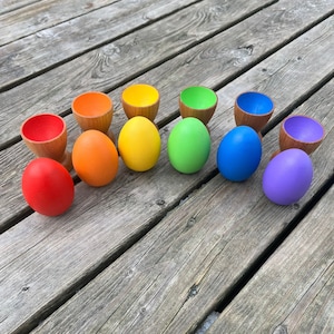 May include: A set of ten wooden egg cups and eggs painted in rainbow colours. The eggs are arranged in a row on a wooden surface.