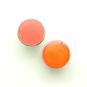 May include: Two round, orange and coral-colored buttons with silver-colored metal rims. The buttons are glossy and appear to be made of enamel or resin. The buttons are set against a white background.