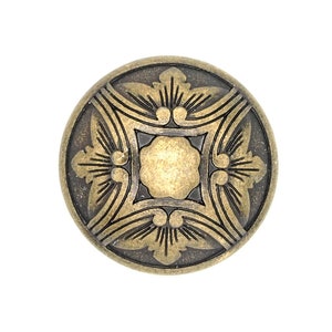 Op de afbeelding: Een ronde, goudkleurige metalen knoop met een decoratief, ingewikkeld ontwerp. De knoop heeft een verhoogd, rond midden en een gedetailleerd, bloemig patroon rond de rand.