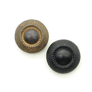 Könnte beinhalten: Zwei runde Knöpfe mit einem erhabenen Zentrum. Ein Knopf ist golden mit einem schwarzen Zentrum und der andere ist schwarz mit einem goldenen Zentrum.