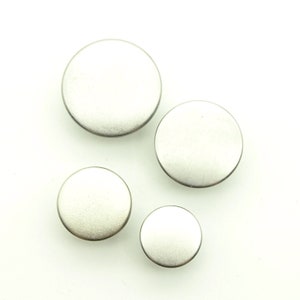 May include: Four silver-coloured, round buttons of varying sizes are arranged on a white background. The buttons have a matte finish and a slightly raised edge, suggesting a metal composition. Sizes range from small to large.
