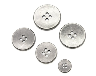 Op de afbeelding: Vijf zilveren metalen knopen met elk vier gaten. De knopen zijn van verschillende grootte en zijn in een halve cirkel gerangschikt.