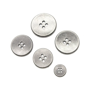 Peut inclure: Cinq boutons en métal argenté avec quatre trous chacun. Les boutons sont de tailles différentes et sont disposés en demi-cercle.