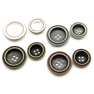 May include: Eight round buttons of varying sizes and finishes. The buttons have a four-hole design and are in colours such as antique brass, silver, and black. Some have a clear or white outer ring.