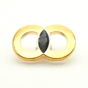 Op de afbeelding: Een goudkleurige broche met een achtvormig ontwerp, met een zwarte, ovaalvormige middensteen. De broche heeft een gepolijste afwerking en een speldsluiting. Dit sieraad is geschikt om een vleugje elegantie aan elke outfit toe te voegen.