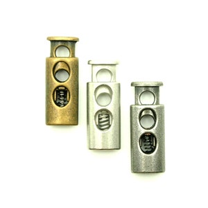 Könnte beinhalten: Drei Metall-Schieber in Gold-, Silber- und Gunmetal-Finish. Jeder Schieber verfügt über einen Federmechanismus und zwei Löcher zum Befestigen von Schnüren oder Bändern.