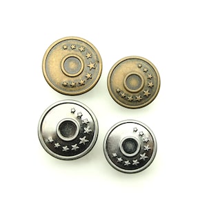 Op de afbeelding: Vier metalen jeansknopen in twee maten. Twee zijn bronskleurig met een sterrenpatroon rond een centrale cirkel. De andere twee zijn zilverkleurig met een vergelijkbaar sterrenontwerp. Alle knopen hebben een licht verhoogd midden.