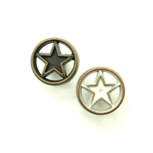 May include: Two metal buttons with a star design. The button on the left has a black star in a bronze circle. The button on the right has a white star in a silver circle with a copper rim.