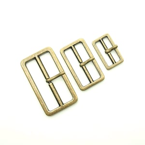 May include: Three antique gold-toned metal belt buckles of different sizes. The buckles are rectangular with a center bar and two side bars.