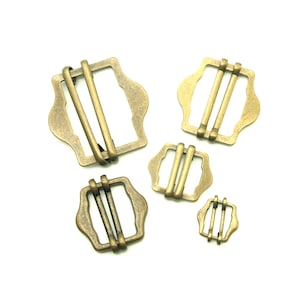 May include: Five antique gold-toned metal buckles of varying sizes. The buckles have a rectangular shape with rounded corners and two parallel bars across the center.