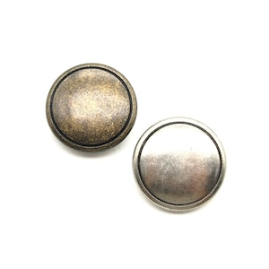 May include: Two round metal buttons. One button has a bronze finish, and the other has a silver finish. Both buttons have a raised edge and a flat centre. The buttons are likely for clothing or craft projects.