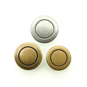 Puede incluir: Tres botones redondos de metal con un borde elevado. Los botones son de plata, latón y latón antiguo.