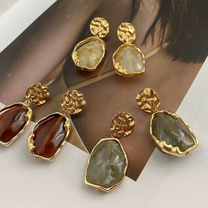 May include: Three pairs of drop earrings featuring gold-toned, hammered-texture circular studs connected to irregular, organic-shaped pendants. The pendants come in translucent cream, deep amber, and muted sage green resin with gold-toned edges.